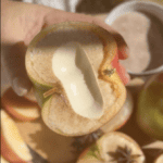 A hand holds a sliced apple with a hollowed-out center filled with cream, dusted with cinnamon.